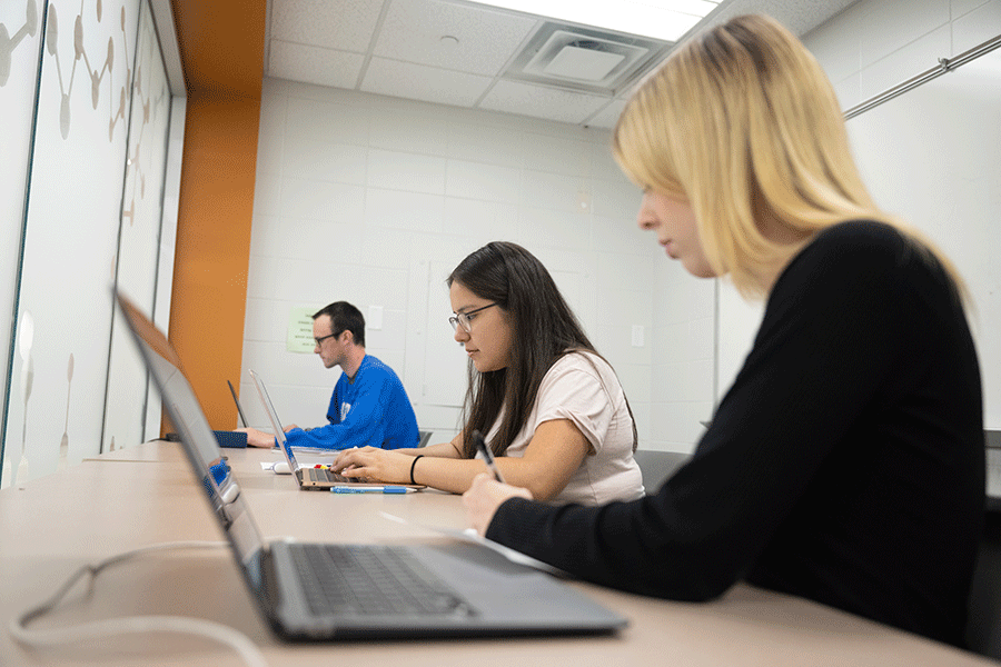 Three students – a blonde woman in a black sweater, a woman with brown hair and wearing a white shirt, and a man with dark hair and glasses wearing a blue sweatshirt, type on their laptops in a well-lit, white-walled classroom.