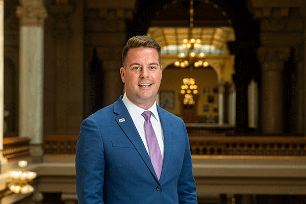 White male with brown hair dressed professionally with suit coat and standing against a railing on the second floor of the Indiana Statehouse in Indianapolis.