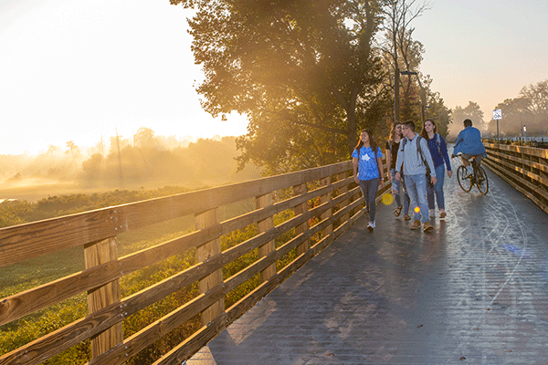 A group of four students walking along a wooden boardwalk that overlooks a scenic open area of marshland in early morning as the sun breaks through the clouds.