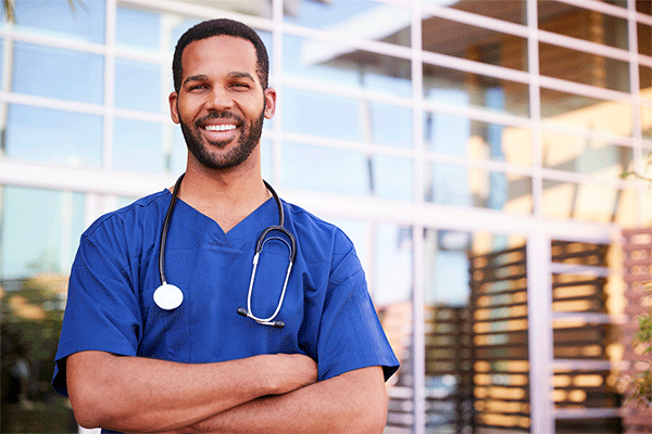 Black male with short hair, mustache and beard in blue nursing scrubs with arms crossed standing outside of a building with glass windows in the background. 