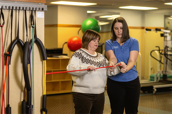 On the left is a white female student with shoulder-length brown hair wearing a blue shirt and black pants. She works with a female client on her right in a physical therapy clinic. The client is a white woman with shoulder-length brown hair wearing a white and brown sweater and black pants. They’re holding a red elastic physical therapy band and other equipment is visible in the background. 