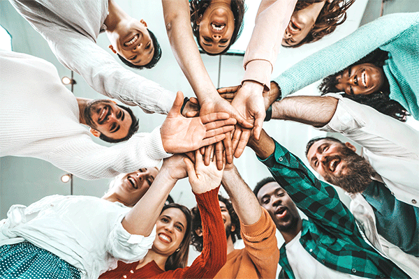 Multicultural group of people stacking hands together in a circle and looking down at the camera. 